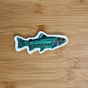 Peut inclure: Écusson brodé représentant un poisson avec un corps turquoise, des détails noirs et un œil orange. Le poisson est bordé de noir et a une bordure blanche. L'écusson est sur une surface en bois.