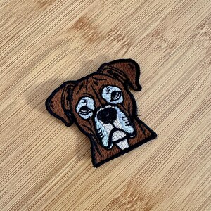 May include: Embroidered patch featuring a Boxer dog's face. The patch has a brown background with black outlining. The dog's face has white, blue, and black details, capturing the breed's distinctive features. Ideal for jackets, bags, or other fabric items.