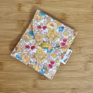 May include: A small floral wallet with a yellow press stud closure. The fabric displays a pattern of yellow, red, and blue flowers on a cream background. The tag reads "Bleu Cerise".