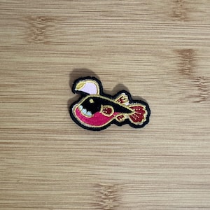May include: Embroidered patch featuring a stylized anglerfish design. The patch has a black outline with gold accents, a pink body, and red fins. The anglerfish has a white lure and a smiling mouth.