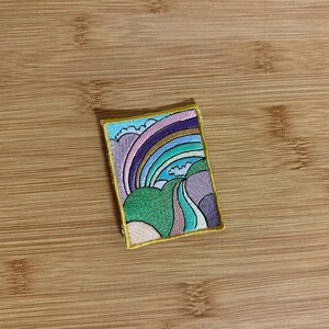 May include: Embroidered patch featuring a colorful rainbow design with shades of purple, blue, green, and yellow. The patch has a gold border and is rectangular in shape. The design includes stylized clouds and rolling hills.