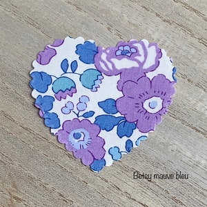 Puede incluir: Un parche de tela en forma de coraz&oacute;n con fondo blanco y un patr&oacute;n floral en tonos de morado y azul. El texto "Betsy mauve bleu" est&aacute; impreso en la parte inferior del parche.