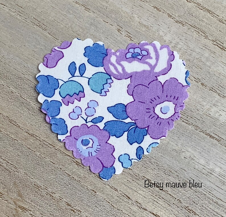Puede incluir: Un parche de tela en forma de coraz&oacute;n con fondo blanco y un patr&oacute;n floral en tonos de morado y azul. El texto "Betsy mauve bleu" est&aacute; impreso en la parte inferior del parche.