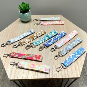 Puede incluir: Una colección de 10 llaveros coloridos con diseños florales. Cada llavero tiene un anillo de metal plateado y una correa de tela con un diseño floral diferente. Los llaveros se muestran en una mesa de madera.