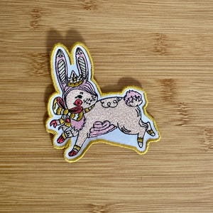 May include: Embroidered patch featuring a whimsical pink bunny wearing a crown and scarf. The bunny is outlined in yellow with a white background. The patch has a playful, cartoonish design, perfect for adding to clothing or accessories.