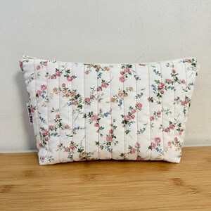 Pode incluir: Uma bolsa de cosméticos acolchoada branca com um padrão floral em rosa, azul e amarelo. A bolsa tem fecho de correr e é costurada verticalmente com linha rosa. A bolsa está sobre uma superfície de madeira.