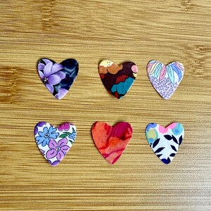 Peut inclure: Six cœurs en tissu avec différents motifs floraux. Les cœurs sont alignés sur une surface en bois. Les cœurs sont de couleur violet, marron, bleu, rose, rouge et blanc.