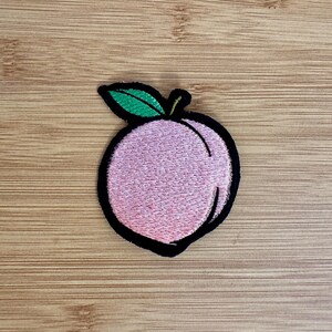 May include: Embroidered patch featuring a pink peach with a green leaf and black outline. The peach is a light pink color with a textured appearance. The patch is likely designed to be ironed or sewn onto fabric.
