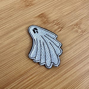 Op de afbeelding: Een wit spookvormig embleem met zwarte omtrek en details. Het embleem is geborduurd met een eenvoudig ontwerp en is waarschijnlijk bedoeld voor toepassing op kleding of accessoires. Het embleem ligt op een houten oppervlak.