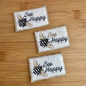 Peut inclure: Trois patchs en feutre blanc avec une bordure de paillettes dorées. Chaque patch présente une abeille brodée en noir et blanc avec le texte "Bee Happy" en broderie noire.