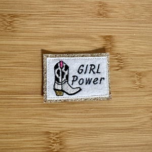 Peut inclure: Patch brodé blanc avec une botte de cow-boy noire et blanche et le texte "GIRL POWER" en noir. Le patch a une bordure dorée pailletée.