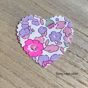Puede incluir: Un parche de tela en forma de coraz&oacute;n con fondo blanco y un patr&oacute;n floral en tonos rosa, morado y verde. El texto "Betsy neon violet" est&aacute; impreso debajo del parche.