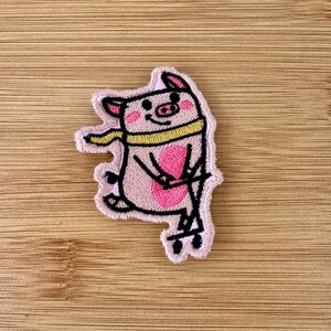 May include: Embroidered patch featuring a pink pig wearing a yellow scarf and riding a skateboard. The pig has a pink heart-shaped belly and black outlines. The patch is on a wooden surface.