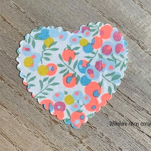 Puede incluir: Un parche de tela en forma de coraz&oacute;n con un fondo blanco y un patr&oacute;n floral en tonos de azul, rosa, amarillo y naranja. La tela tiene un borde festoneado y el texto "Wiltshire neon coral" est&aacute; impreso debajo del parche.