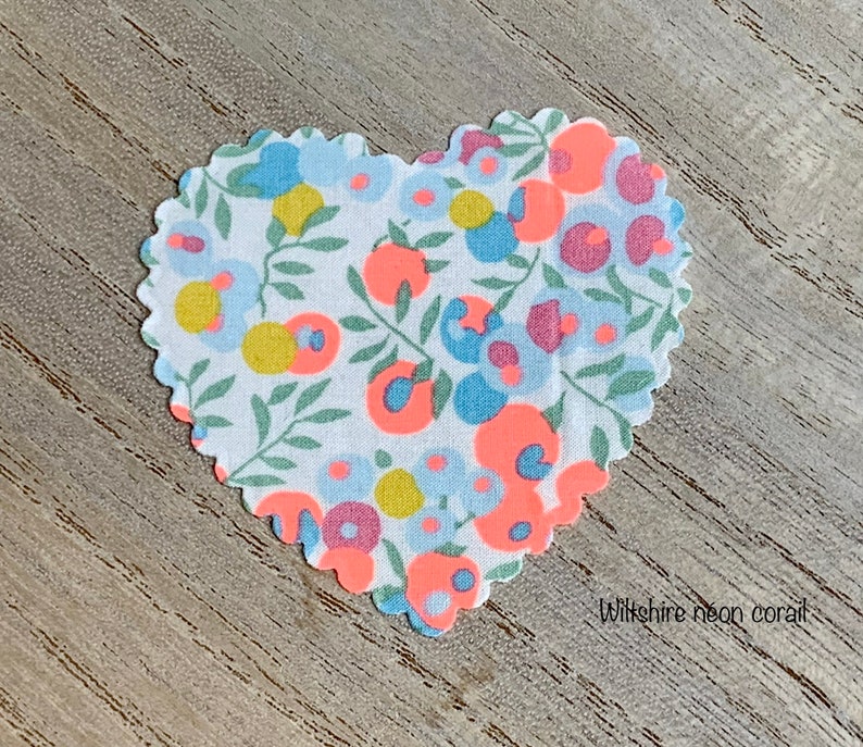 Puede incluir: Un parche de tela en forma de coraz&oacute;n con un fondo blanco y un patr&oacute;n floral en tonos de azul, rosa, amarillo y naranja. La tela tiene un borde festoneado y el texto "Wiltshire neon coral" est&aacute; impreso debajo del parche.