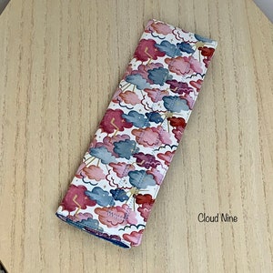 Peut inclure: Un couvre-ceinture de sécurité en tissu blanc avec un motif de nuages roses, bleus et dorés. Le texte "Cloud Nine" est imprimé dans le coin inférieur droit de l'image.
