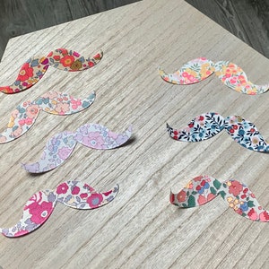 Könnte beinhalten: Sieben Papier-Ausschnitte von Schnurrbärten in verschiedenen Blumenmustern. Die Schnurrbärte befinden sich auf einer hellbraunen Holzoberfläche.