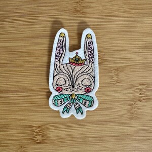 May include: Embroidered patch featuring a stylized rabbit with a crown, closed eyes, and a bow. The patch has a white border, with pink, green, and gold accents. The rabbit is tan with black details.