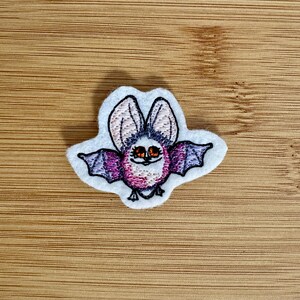 May include: A white felt bat patch with a cartoon design. The bat has large ears, a pink body, and purple wings. The bat's eyes are orange, and the design is outlined in black.