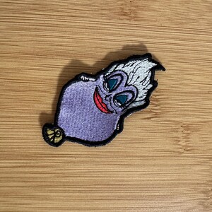 May include: A purple embroidered patch featuring a cartoon character with white hair, teal eyes, and a red smile. The patch has a black border and a yellow and gold detail at the bottom. The patch is on a wooden surface.