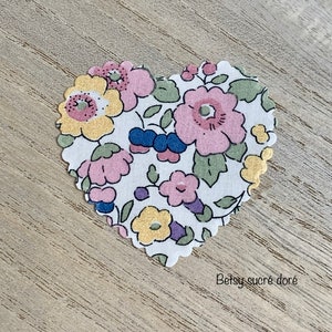 Puede incluir: Un parche de tela blanco en forma de coraz&oacute;n con un patr&oacute;n floral en tonos de rosa, amarillo, verde y azul. La tela tiene un borde festoneado. El texto "Betsy sucre dor&eacute;" es visible debajo del parche.
