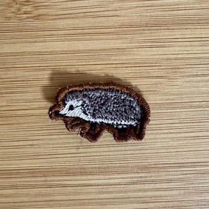 May include: Embroidered patch featuring a hedgehog design. The patch has a brown border, gray and white body, and a detailed face. The patch is placed on a wooden surface.