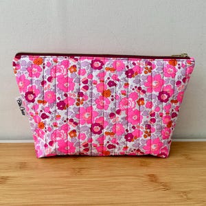 Pode incluir: Uma bolsa acolchoada com fecho de correr apresentando um padrão floral vibrante em tons de rosa choque, magenta e laranja sobre um fundo branco. O saco retangular tem linhas de costura verticais e uma pequena etiqueta de tecido branco na lateral.