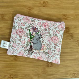 Puede incluir: Pequeño estuche con cremallera con un estampado floral rosa y blanco. El estuche presenta un ratón gris bordado sosteniendo una flor. Una pequeña etiqueta con el texto "Blan Cerise" está adjunta al costado.
