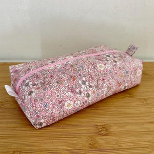 Peut inclure: Trousse rectangulaire avec un motif floral rose. La trousse a une fermeture éclair rose et une petite boucle en tissu à une extrémité. Le motif floral comprend des fleurs blanches, bleues et jaunes sur fond rose. La trousse est en tissu.