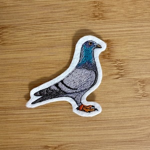 May include: Embroidered patch of a pigeon with a blue head and neck, grey body, and orange feet. The patch is outlined in white. The background is a wooden surface.