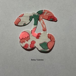 Peut inclure: Une cerise en tissu blanc avec un imprimé floral rose et vert. La cerise est découpée et a une tige avec une feuille. Le texte "Betsy Tuileries" est sous la cerise.