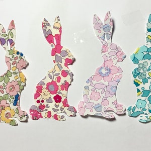 May include: Four paper cut-outs of bunnies in different colours and patterns. The bunnies are all facing the right side of the image. The bunnies are all in a sitting position. The bunnies are all made of paper with a floral pattern. The bunnies are all in different colours: pink, blue, green, and yellow.