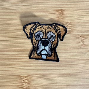 May include: An embroidered patch featuring the head of a boxer dog with tan fur, a black nose, and white markings around the eyes and muzzle. The design is outlined in black thread, capturing the distinct facial features and floppy ears of the breed.