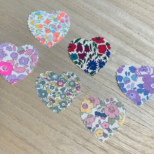 May include: Six heart-shaped fabric patches with floral patterns in shades of pink, blue, and white. The patches are arranged in a semi-circle on a wooden surface.