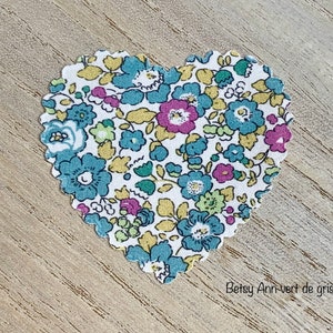 Puede incluir: Un parche de tela en forma de coraz&oacute;n con un patr&oacute;n floral en tonos de azul, amarillo, rosa y verde. La tela tiene un fondo blanco y el texto "Betsy Ann-vert de gris" est&aacute; impreso debajo del coraz&oacute;n.