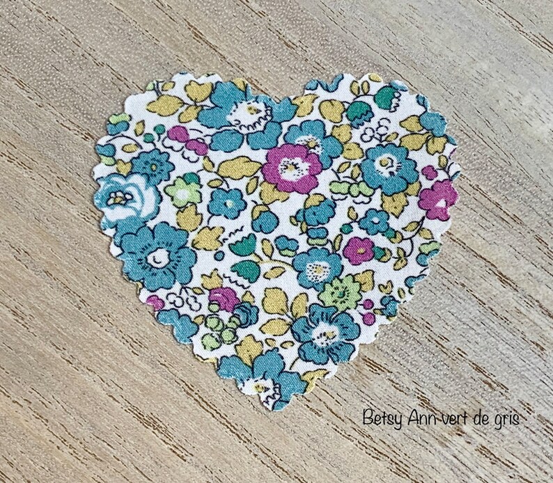 Puede incluir: Un parche de tela en forma de coraz&oacute;n con un patr&oacute;n floral en tonos de azul, amarillo, rosa y verde. La tela tiene un fondo blanco y el texto "Betsy Ann-vert de gris" est&aacute; impreso debajo del coraz&oacute;n.