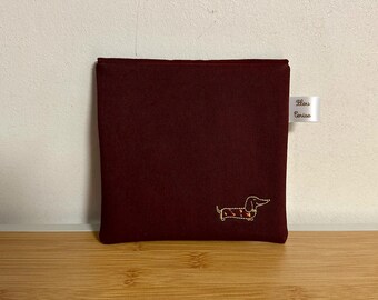 May include: A burgundy fabric pouch with a gold embroidered dachshund design. The pouch has a white label with the text "Fleur Denise" on it.