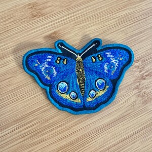 May include: Embroidered butterfly patch with a vibrant blue design. The patch features a detailed pattern with yellow and white accents, outlined in black. The butterfly has a detailed body and wing patterns.