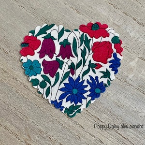 Puede incluir: Un parche de tela en forma de coraz&oacute;n con fondo blanco y un patr&oacute;n floral. El patr&oacute;n incluye amapolas rojas, campanillas moradas, flores azules y hojas verdes. El texto "Poppy Daisy bleu canard" est&aacute; impreso debajo del parche.