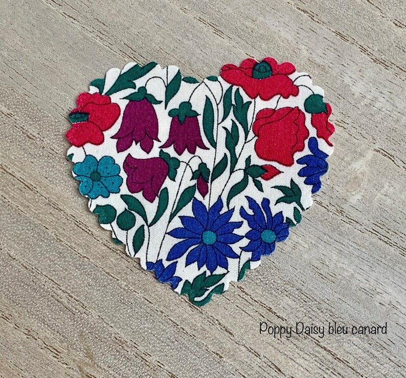 Puede incluir: Un parche de tela en forma de coraz&oacute;n con fondo blanco y un patr&oacute;n floral. El patr&oacute;n incluye amapolas rojas, campanillas moradas, flores azules y hojas verdes. El texto "Poppy Daisy bleu canard" est&aacute; impreso debajo del parche.