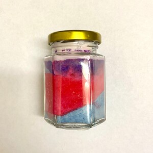 May include: A glass jar with a gold lid filled with layered blue, red, and purple bath salts.