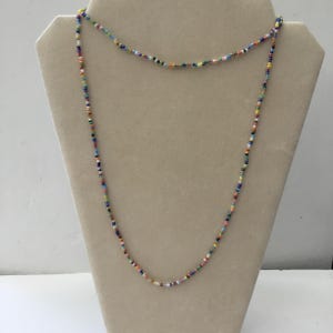 May include: A long beaded necklace featuring a variety of small, colourful beads. The beads are in shades of red, blue, green, yellow, and white. The necklace is displayed on a beige mannequin.