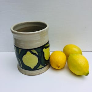 May include: A ceramic vase with a beige top and a green and yellow lemon pattern. Three yellow lemons are arranged to the right of the vase. The vase is cylindrical with a wide opening and a slightly flared base.