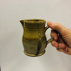 May include: A small, brown ceramic jug with a speckled glaze. The jug has a rounded body and a small handle.