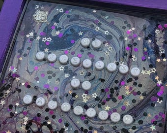 May include: A framed artwork with a purple border. The artwork features a swirling, abstract design in shades of blue, purple, and white, with scattered glitter and star-shaped confetti. The words "BE EXCELLENT TO EACH OTHER" are spelled out in white beads.