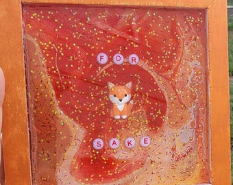 May include: Orange framed artwork with a resin background in shades of red and orange, with gold glitter accents. A small fox figurine is centered, with the words "FOR SAKE" spelled out in letter beads.