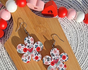 May include: A pair of flower-shaped earrings with a floral pattern of red, pink, and green on a white background. Each earring has a small red heart in the center. The earrings are displayed on a wooden cutting board with decorative beads.