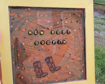 May include: A framed artwork with a gold border. The artwork features the words "NEW BOOT GOOFIN" spelled out in black letter beads. Below the text are two brown cowboy boots with star designs. The background is a mix of orange and brown.
