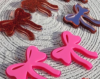 May include: A pair of pink bow earrings and a pair of red glitter bow earrings. The earrings are on a white background with red and pink hearts.