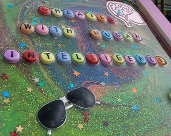 May include: A framed artwork with the words "CREATED WITH HUMAN INTELLIGENCE" spelled out in colorful letters. The artwork features a green and glittery background, star-shaped confetti, and a pair of sunglasses.